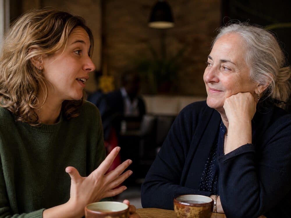Two people of different generations sitting across a small table in conversation, making eye contact, one smiling as the other speaks